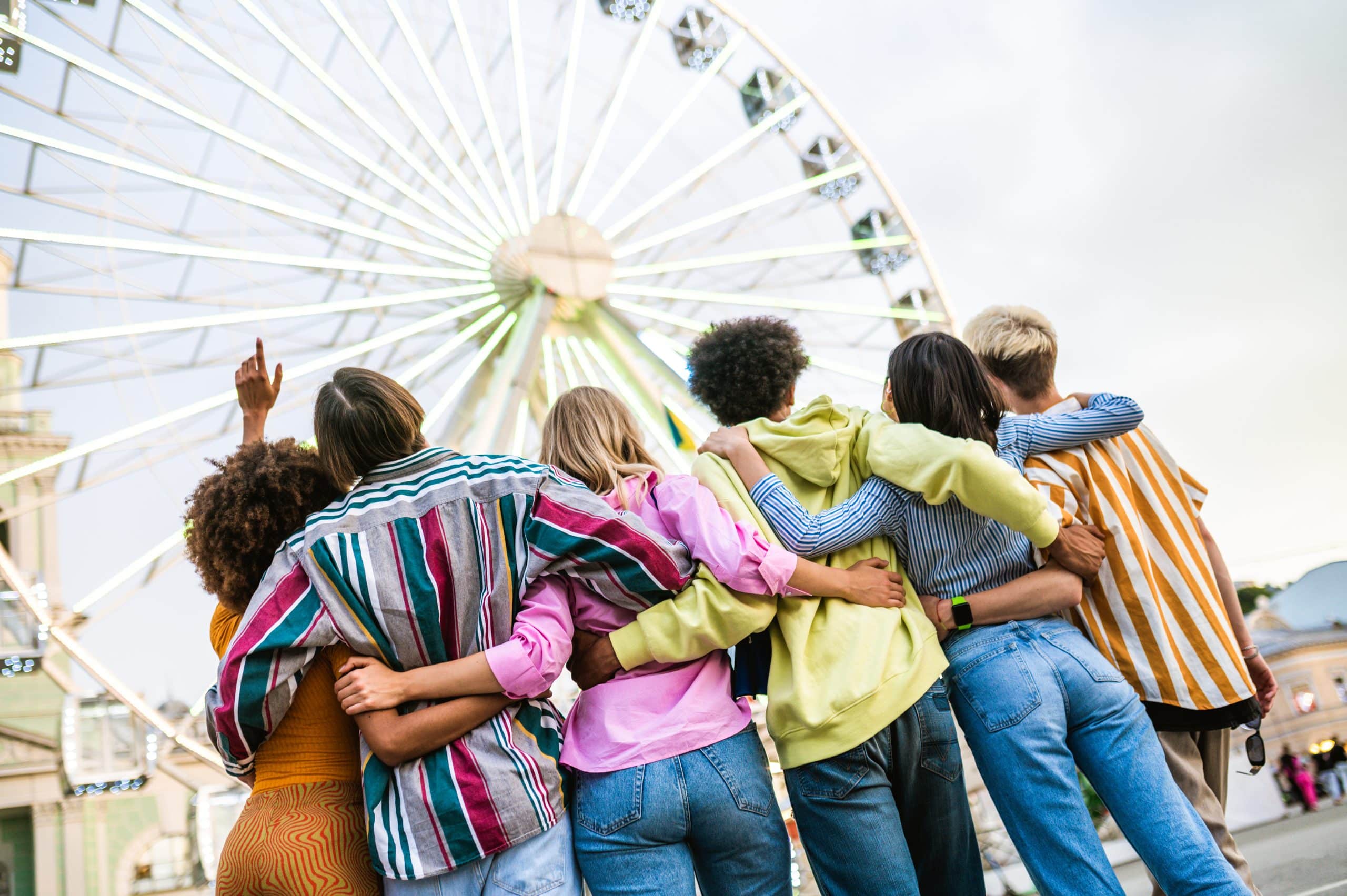 Youth group bus transporting Group of diverse young friends enjoying together at a fair in St Paul, standing arm in arm in front of a large Ferris wheel, representing fun outdoor events and multicultural gatherings. Get a youth group bus for your next outing!